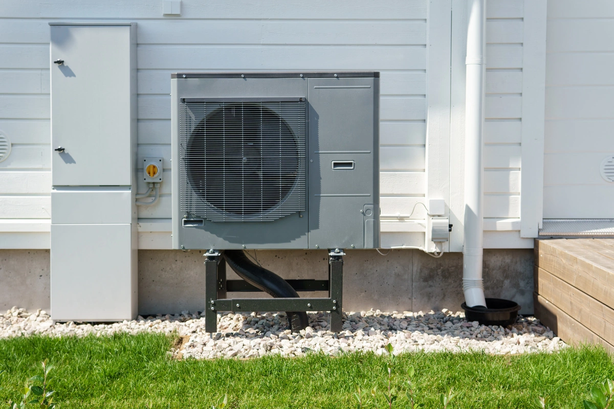 Pompe à chaleur air/eau grise installée à l'extérieur d'une maison blanche, sur une base de gravier.